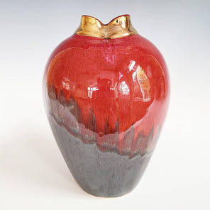 A red and black glazed vessel with a short gold opening sits on a white surface.