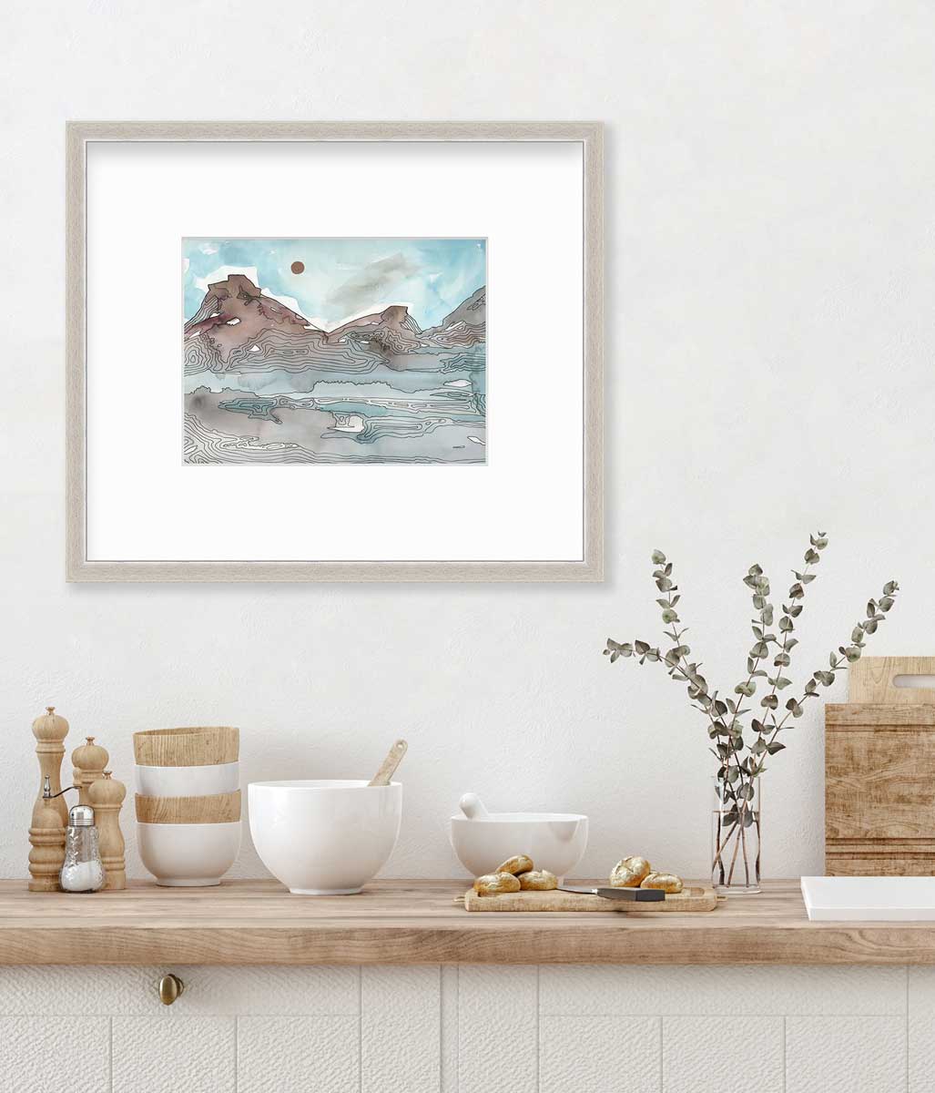 A light blue, brown and black abstract landscape illustration print in a silver frame with a mat hangs on a white wall above a butcher block countertop with bowls, a cutting board and a clear glass vase with eucalyptus stems.