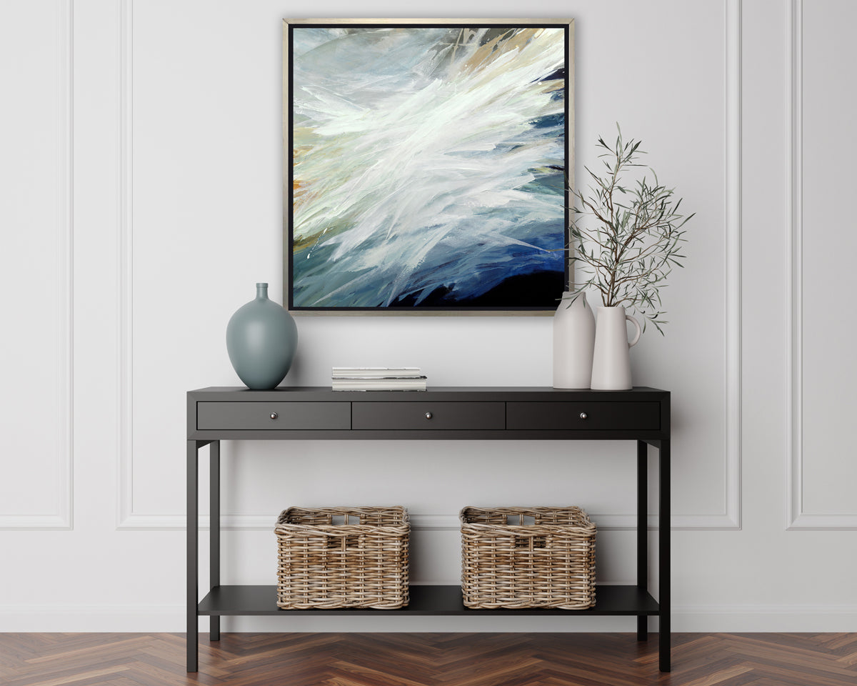 A blue, teal, beige and white abstract print in a silver floater frame hangs on a white wall above a black console table with a muted teal vase, a stack of books, two white vases with greenery stems and two woven baskets.