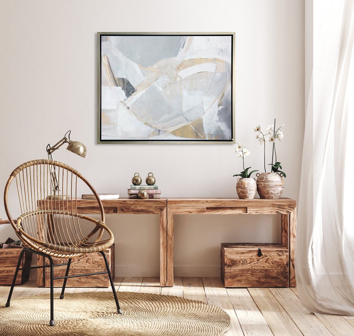 A light grey, beige and white abstract print in a silver floater frame hangs on a white wall above a wood desk with a stack of books, two potted white orchids and a chrome adjustable desk lamp.