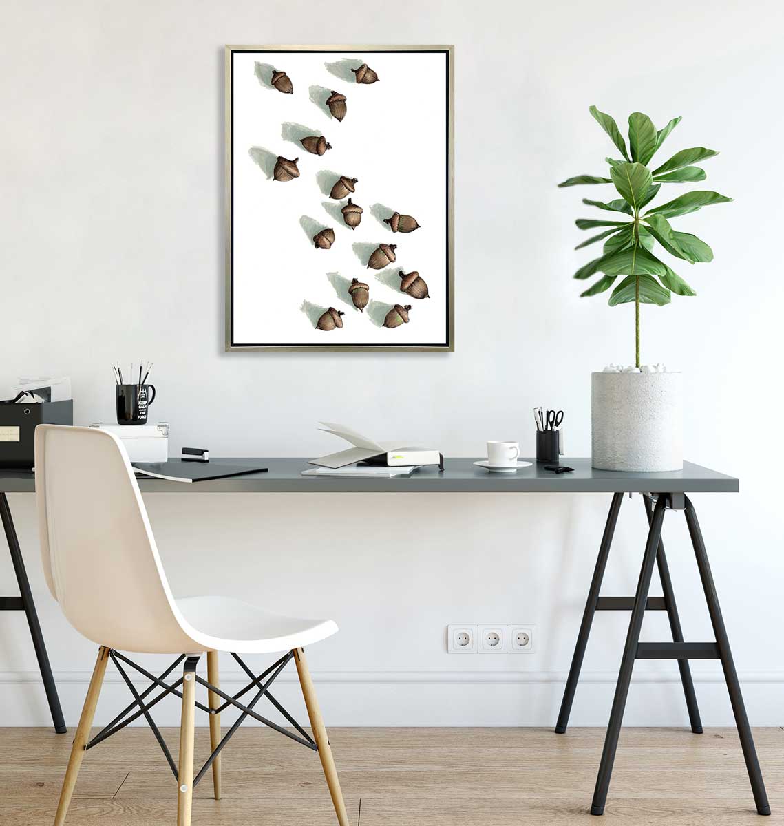A print of several brown acorns with long shadows on a white background in a white silver floater hang on a white wall above a grey desk with several books, a black container with pencils and utensils and a potted tree.