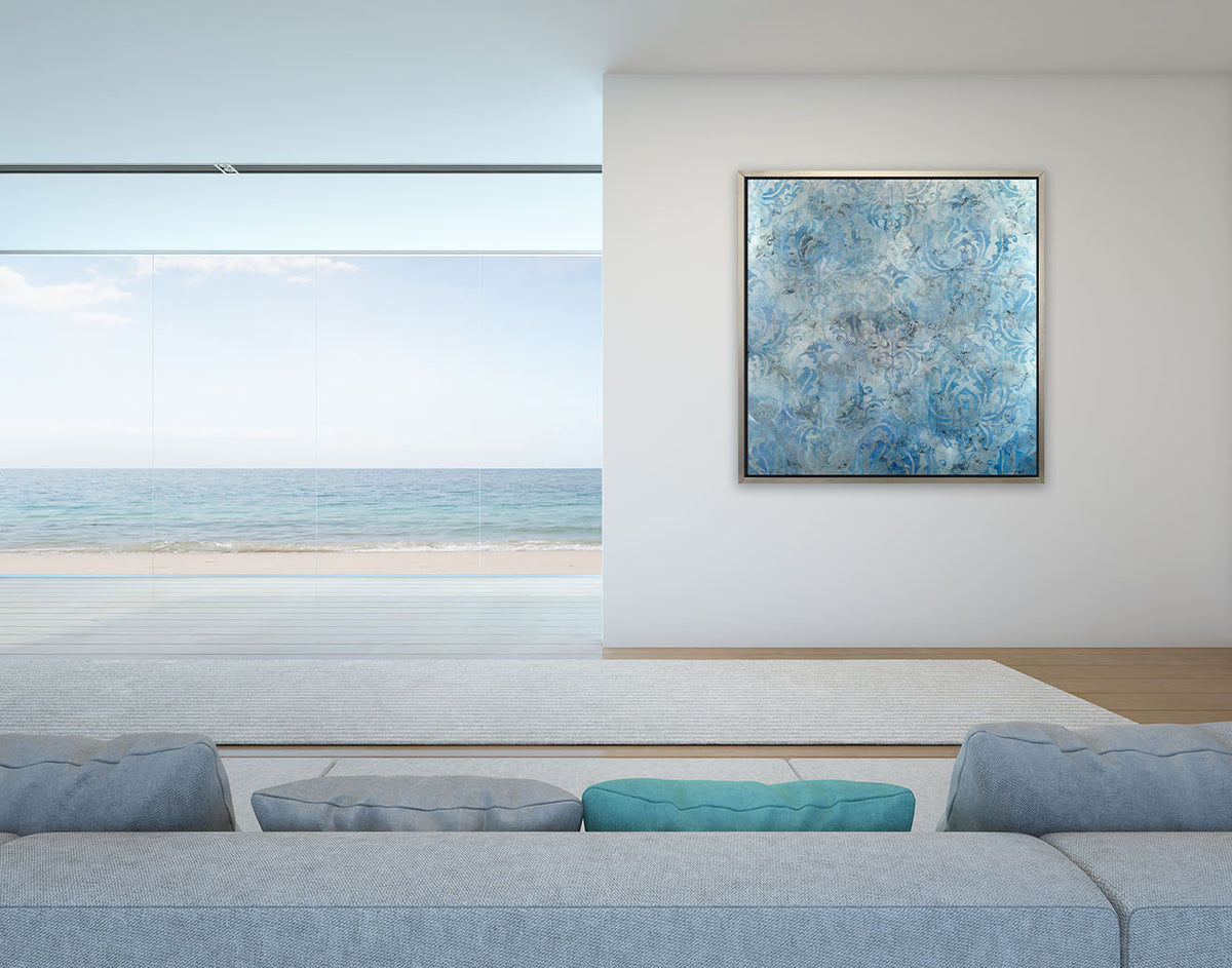 A dark blue, light blue and silver abstract print in a silver floater frame hangs on a white wall across from a long light denim sofa with matching pillows and next to floor to ceiling windows showing a beach.