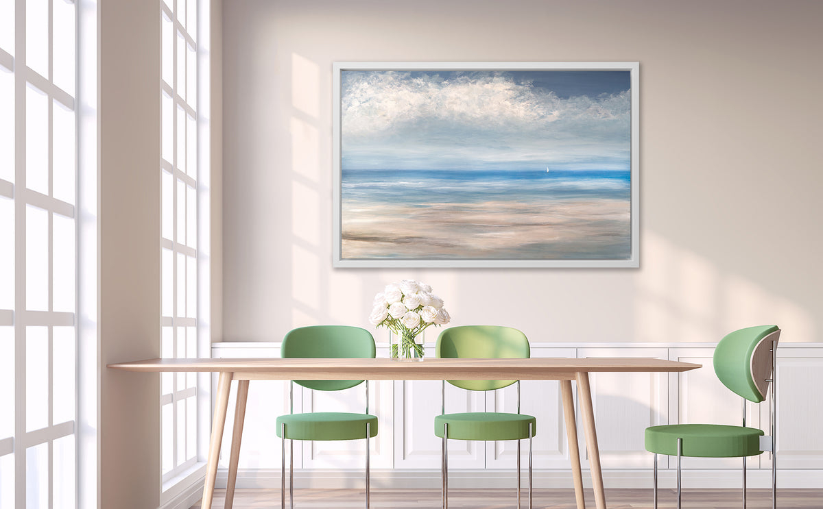 A blue, white and beige seascape print in a white floater frame hangs on a white wall above a wood table with a clear glass vase with a bouquet of white roses and three green chairs surrounding the table.