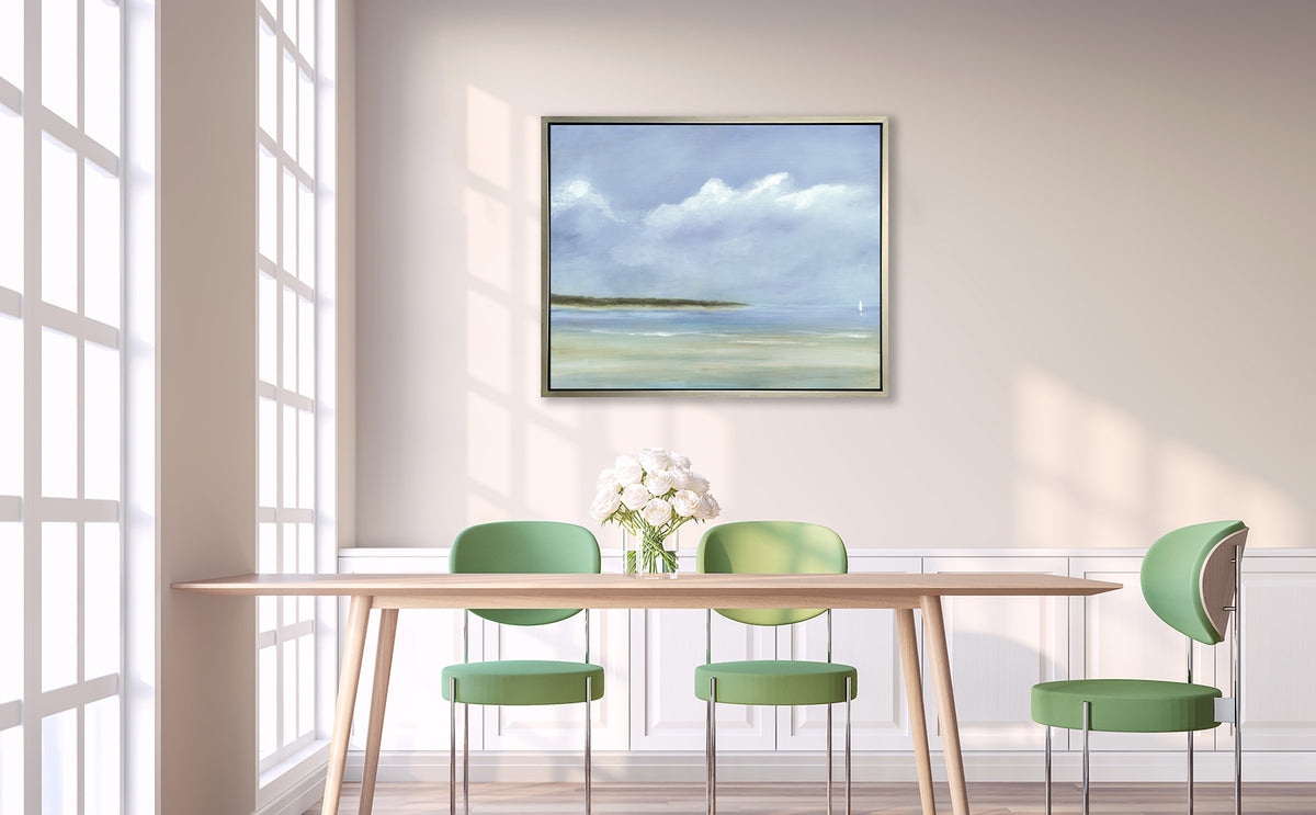 A blue, white and green seascape print in a silver floater frame hangs on a white wall above a wood table with a clear glass vase with a bouquet of white roses and three green chairs surround the table.
