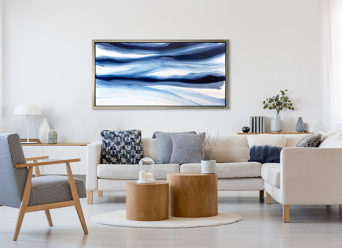 A dark blue and white abstract print in a silver floater frame hangs on a white wall above a white sectional sofa with pillows, two wood cylindrical coffee tables and a blue and white checkered chair.
