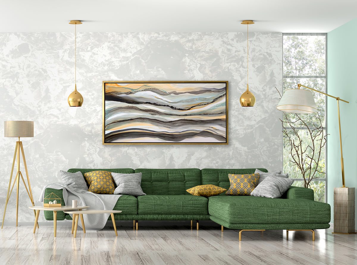 A black, white, orange and teal abstract print in a gold floater frame hangs on a white marble wall above a green sectional sofa with yellow and grey pillows, a grey throw blanket and a two pendant lights.