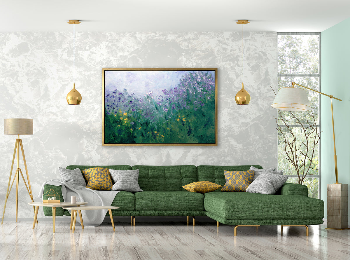 A blue, purple and green abstract print in a gold floater frame hangs on a white marble wall above a green sectional sofa with gold and white pillows, a white throw blanket and two floor lamps on each side.