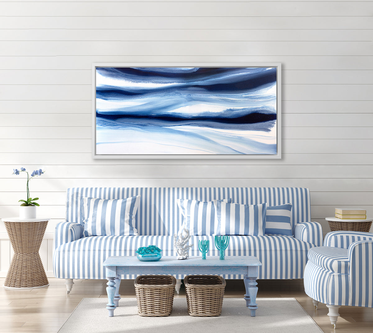 A dark blue and white abstract print in a white floater frame hangs on a white wall above a blue and white striped sofa, a matching chair and a light blue rectangular coffee table.