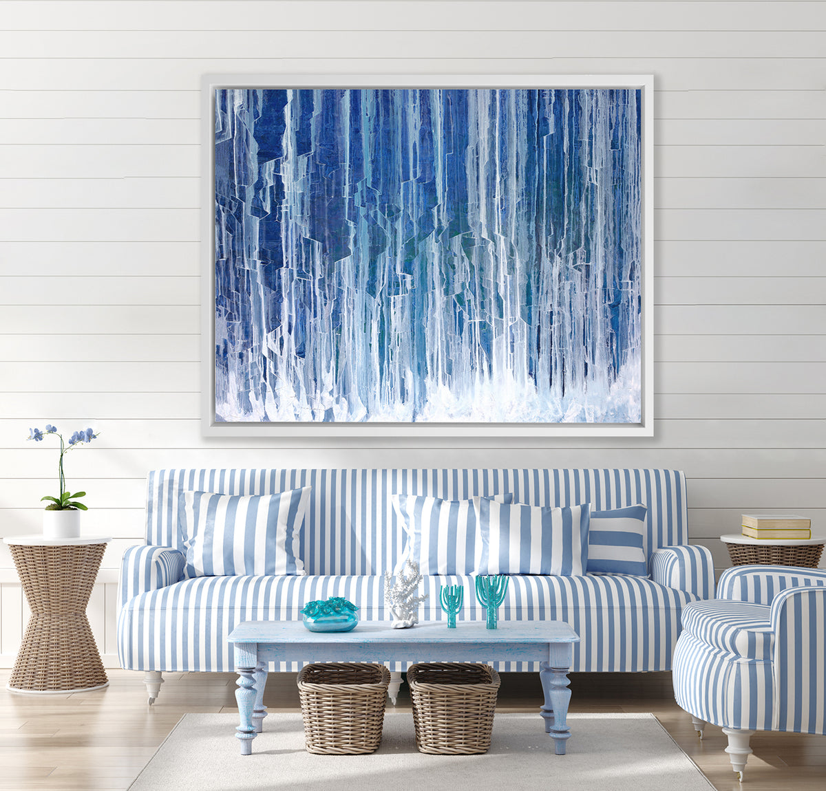 A blue and white abstract print in a white floater frame hangs on a white wall above a blue and white striped sofa, a matching armchair and a blue coffee table.