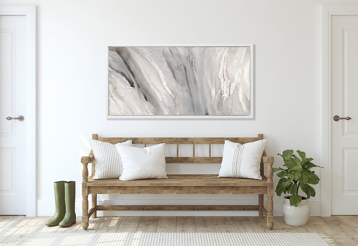 A beige, white and gold abstract print in a white floater frame hangs on a white wall above a wood bench with neutral pillows, a small potted floor plant and a pair of green rubber boots.