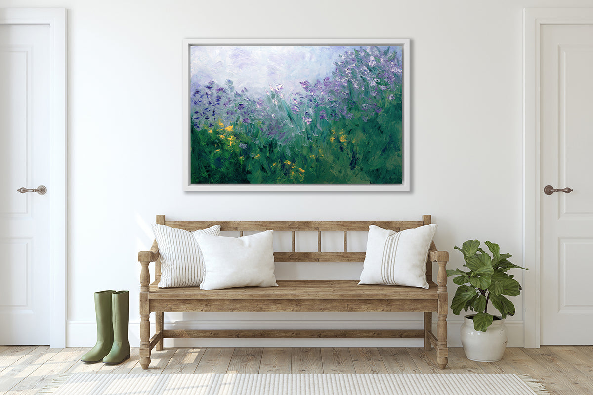 A blue, purple and green abstract print in a white floater frame hangs on a white wall above an armed wood bench with white pillows, a small potted plant and a pair of green rubber boots.