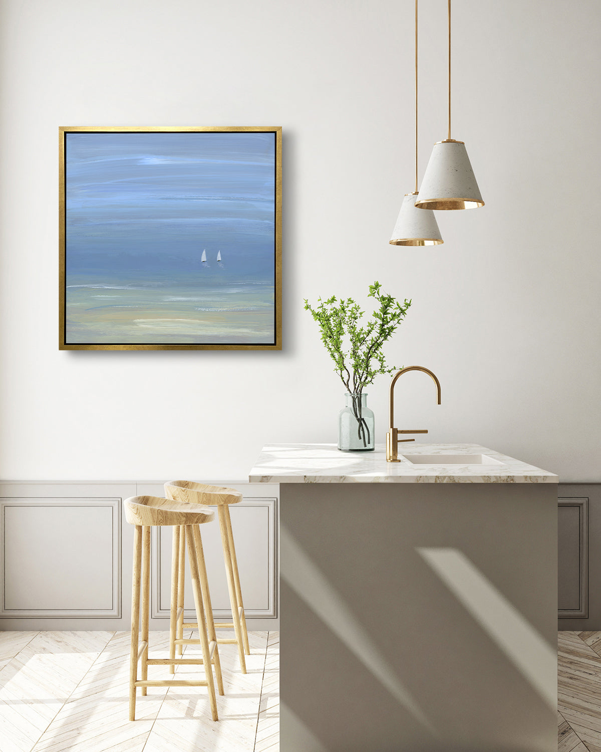 A blue, beige and white abstract seascape print with two small sailboats in a gold floater frame hangs on a white wall above a white marble kitchen island with a sink and a vase with greenery and two wood counter stools.