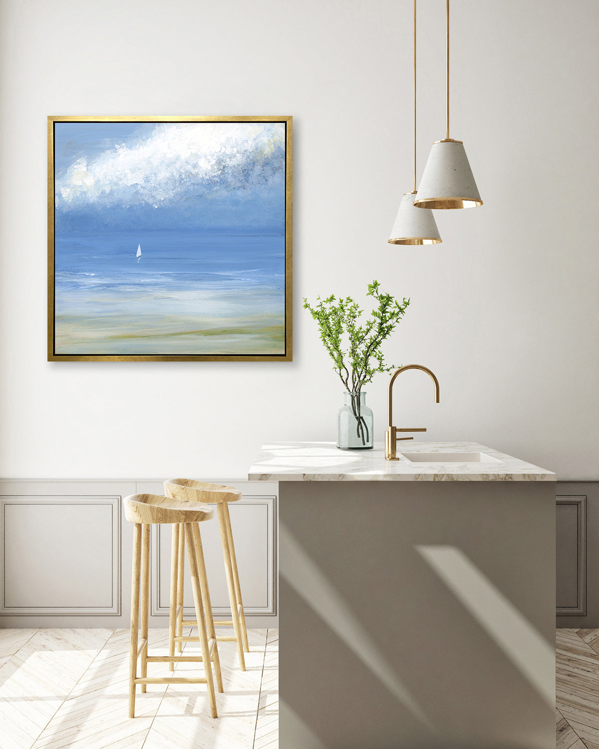 A blue, light green and white abstract seascape print with a small sailboat in a gold floater frame hangs on a white wall above a white marble kitchen island with a sink and a clear vase with greenery stems and two wood counter stools.