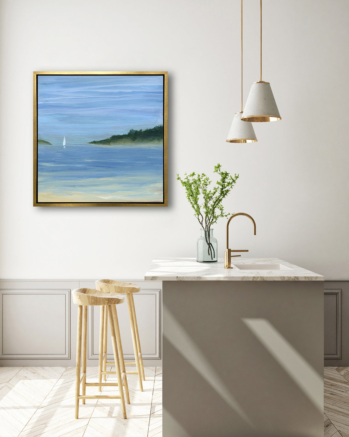 A blue, green and white abstract seascape print with a small sailboat in a gold floater frame hangs on a white wall above a white marble kitchen island with a sink and a clear glass with greenery stems and two wood counter stools.