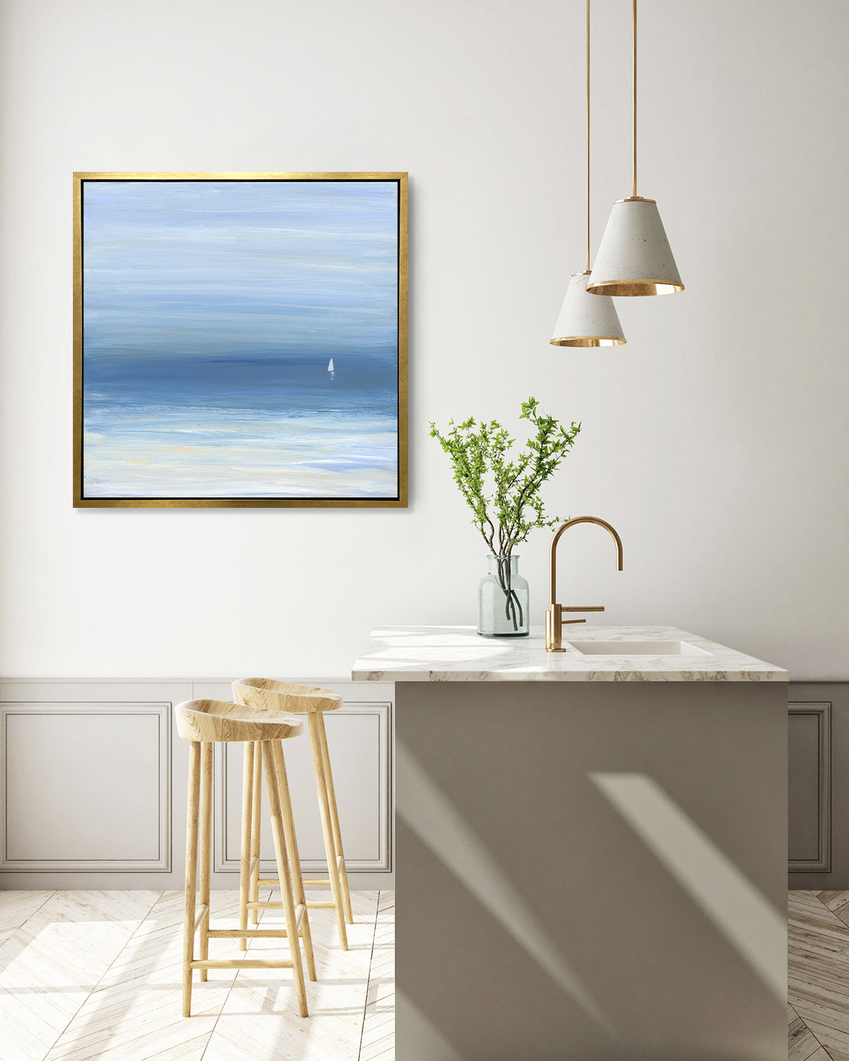 A blue, beige and white abstract seascape print with a small sailboat in a gold floater frame hangs on a white wall above a white marble kitchen island with a sink and a clear vase with greenery stems and two wood counter stools.