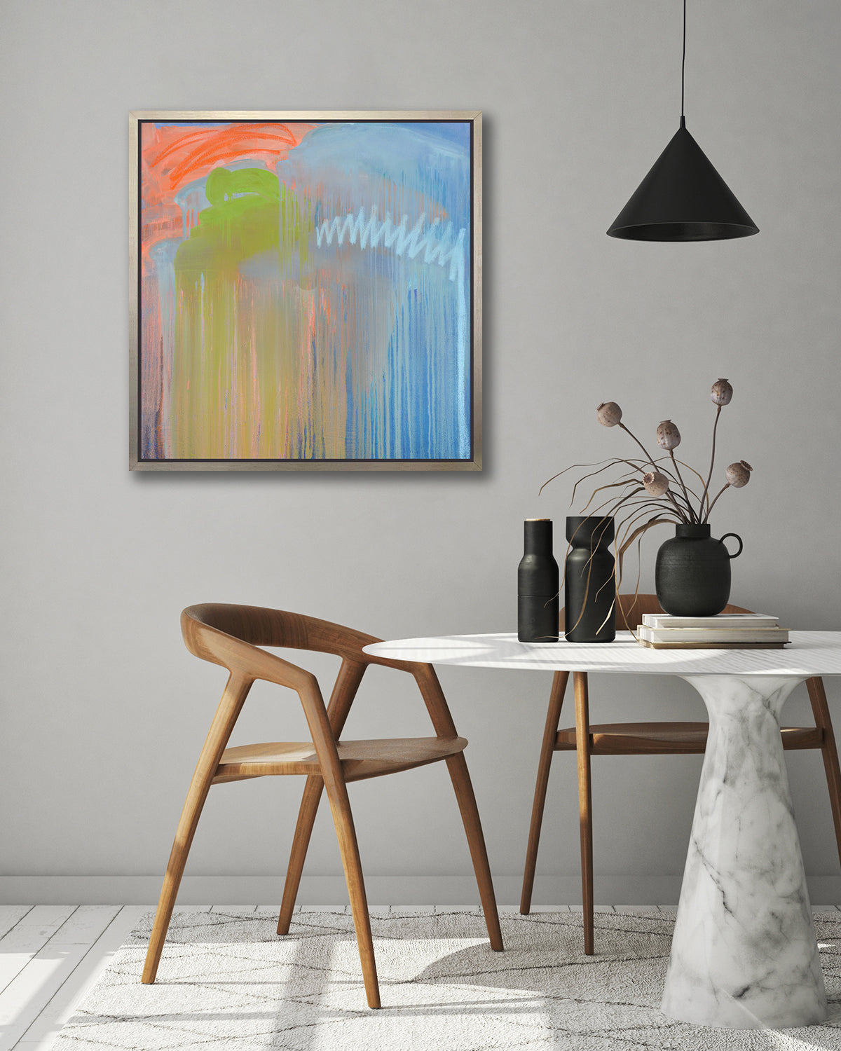 A blue, orange and green abstract print in a silver floater frame hangs on a white wall above two wood chairs and a round white marble pedestal table with black vases and a stack of books. 