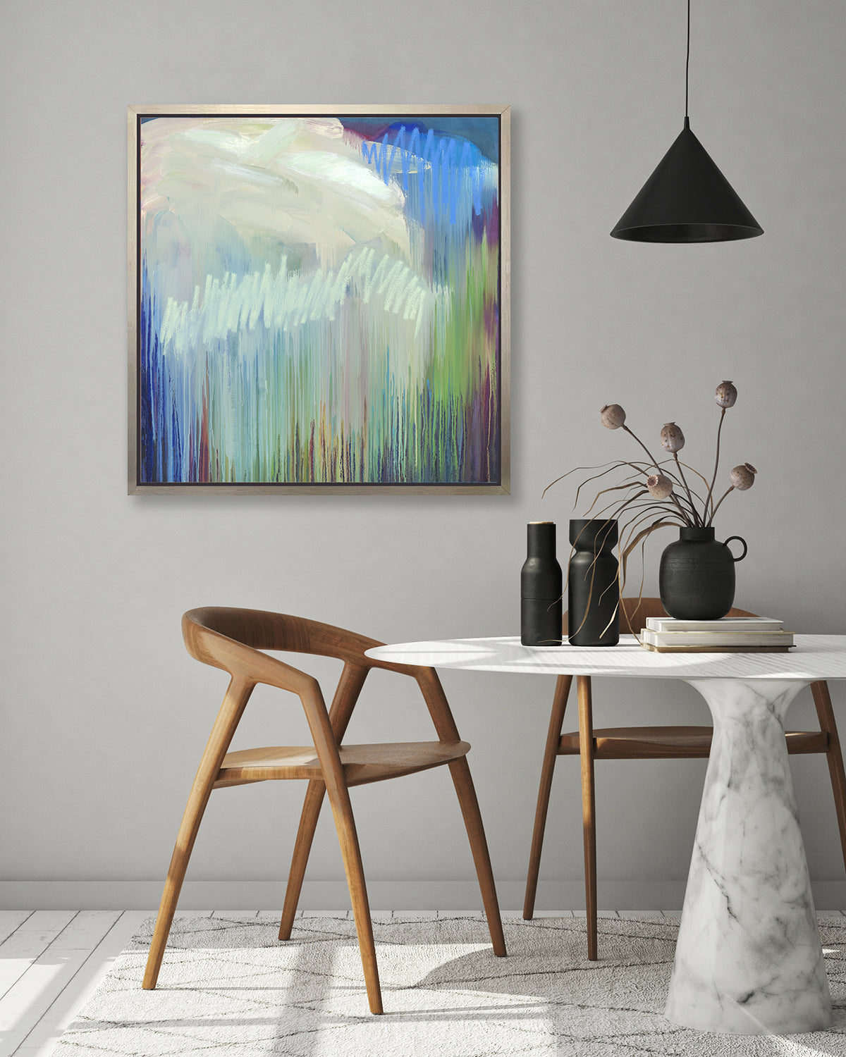 A multicolored abstract print in a silver floater frame hangs on a white wall above two wood chairs and a white marble table with black vases and stack of books.