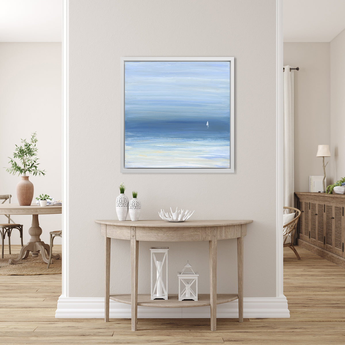 A blue, beige and white abstract seascape print with a small sailboat in a white floater frame hangs on a white wall above a raw wood half circle console table with two white vases, a white decorative bowl and two white lanterns. 