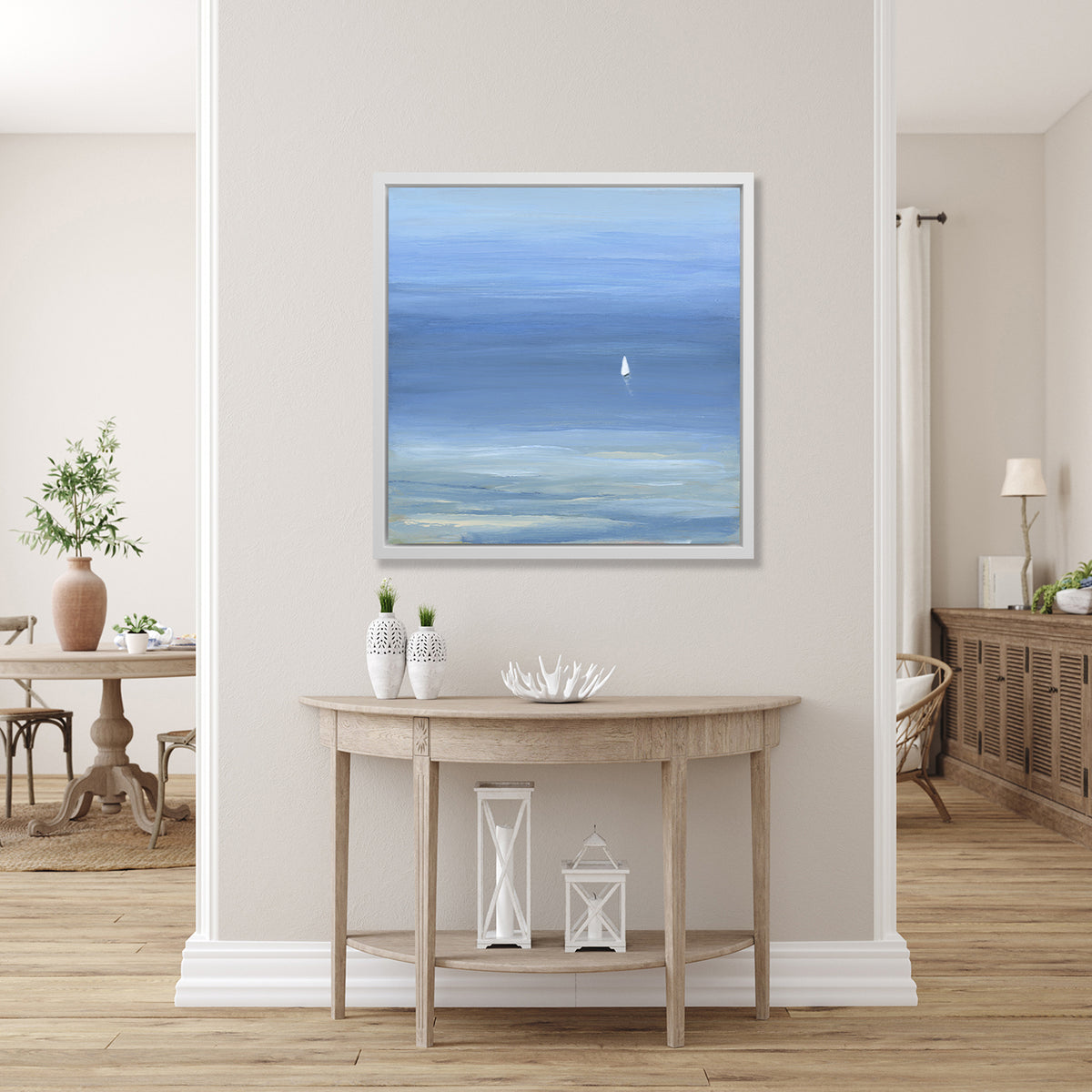 A blue, light green and white abstract seascape print with a small sailboat in a white floater frame hangs on a white wall above a raw wood half circle console table with two white vases, a white decorative bowl and two white lanterns.