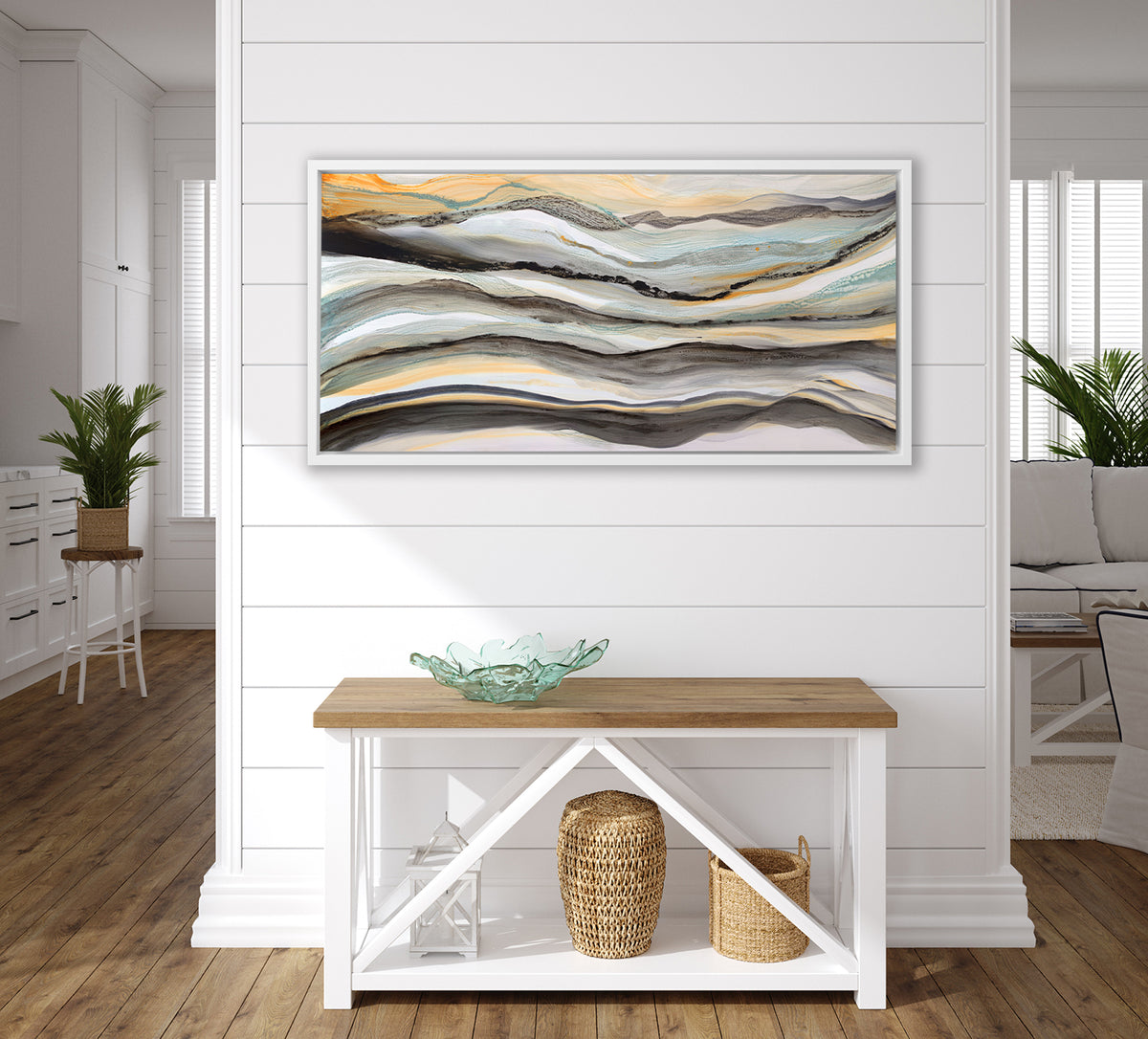 A black, white, orange and teal abstract print in a white floater frame hangs on a white wall above a wood console table with a teal decorative bowl, a white lantern and two woven baskets.