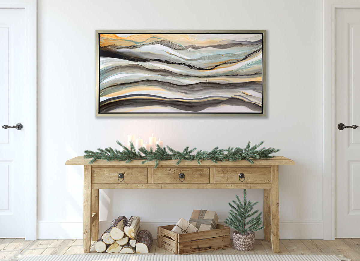 A black, white, orange and teal abstract print in a silver floater frame hangs on a white wall above a wood console table with pine boughs.