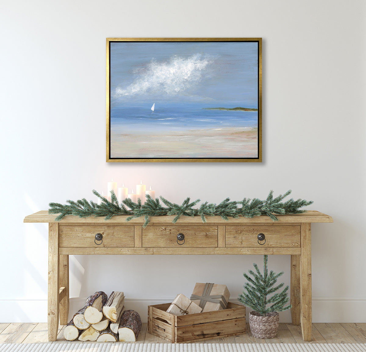 A blue, beige and white abstract seascape print with a small sailboat in a gold frame hangs on a white wall above a wood console table with tree boughs and candles and underneath a pile of a small logs, a palette box with wrapped gifts and a small potted tree.