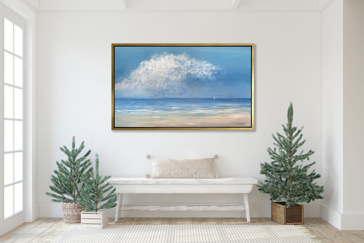 An impressionistic seascape canvas print in a gold floater frame hanging on a white wall above a whitewashed bench with a ivory lumbar pillow and three potted miniature pine trees.