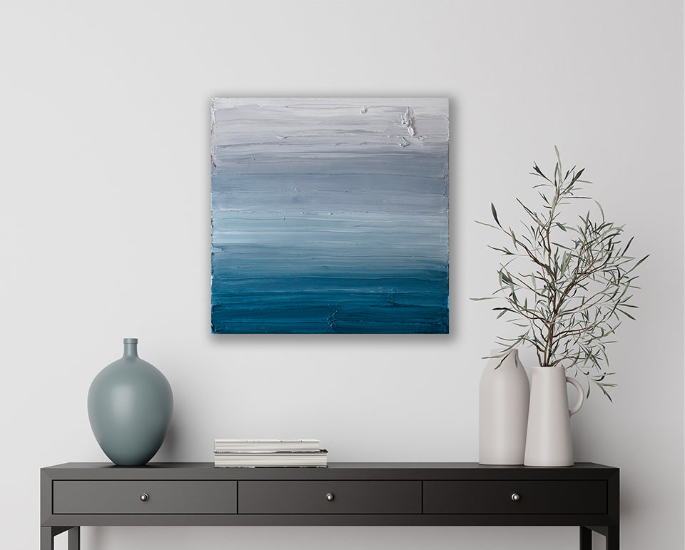 A white, light grey and tonal blues abstract textured painting by Teodora Guererra hangs on a white wall above a dark console table with a stacks of books, a aqua blue tonal vase. On the right side 2 white vases hold an arrangement of greens.