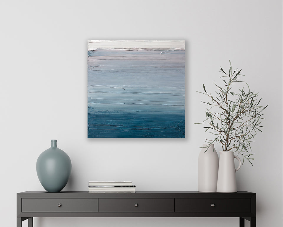 A white, light grey and tonal blues abstract textured painting by Teodora Guererra hangs on a white wall above a dark console table with a stacks of books, a aqua blue tonal vase. On the right side 2 white vases hold an arrangement of greens.
