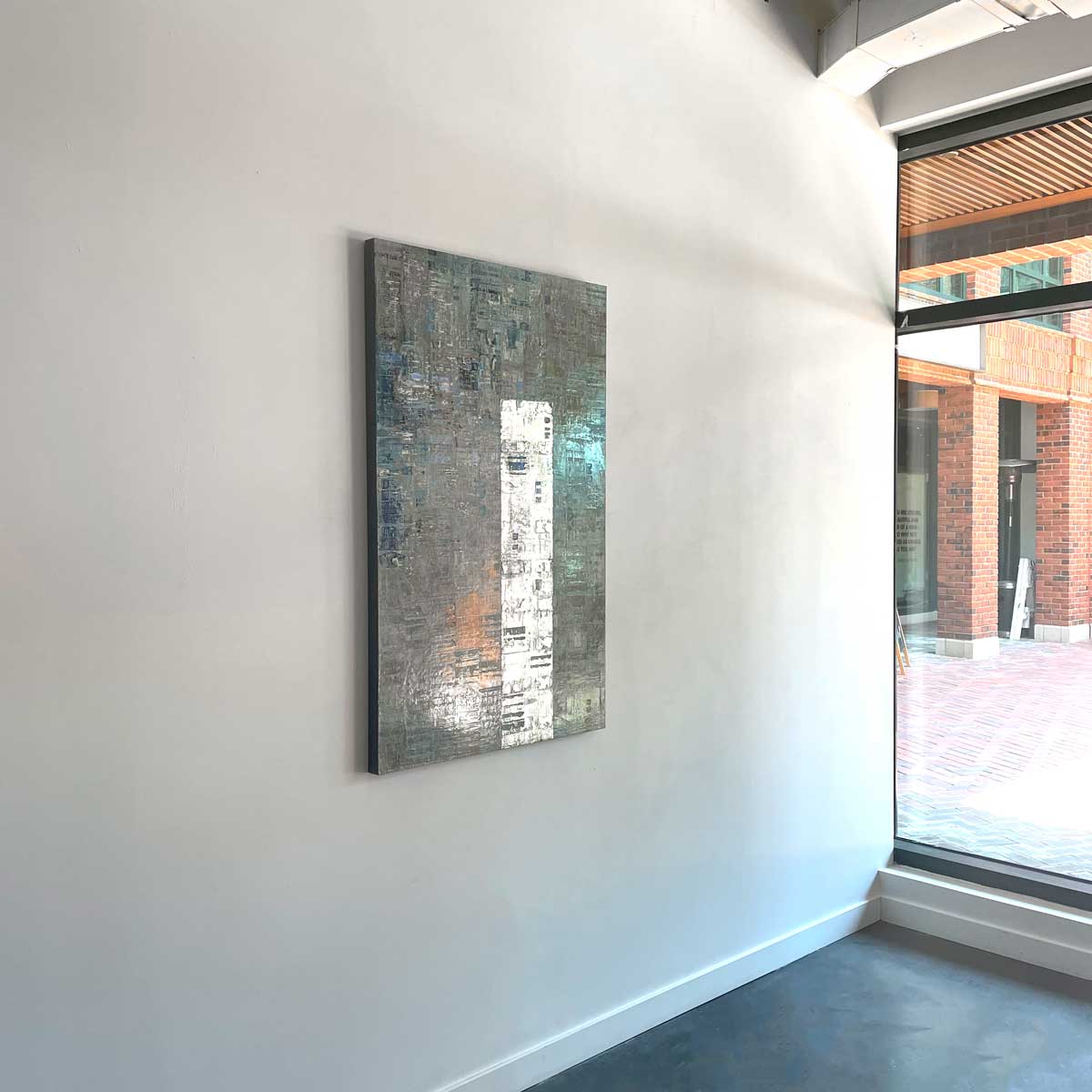 A blue and grey abstract painting with a silver leaf detail by Ned Martin hangs on a white wall with a view from the left. 