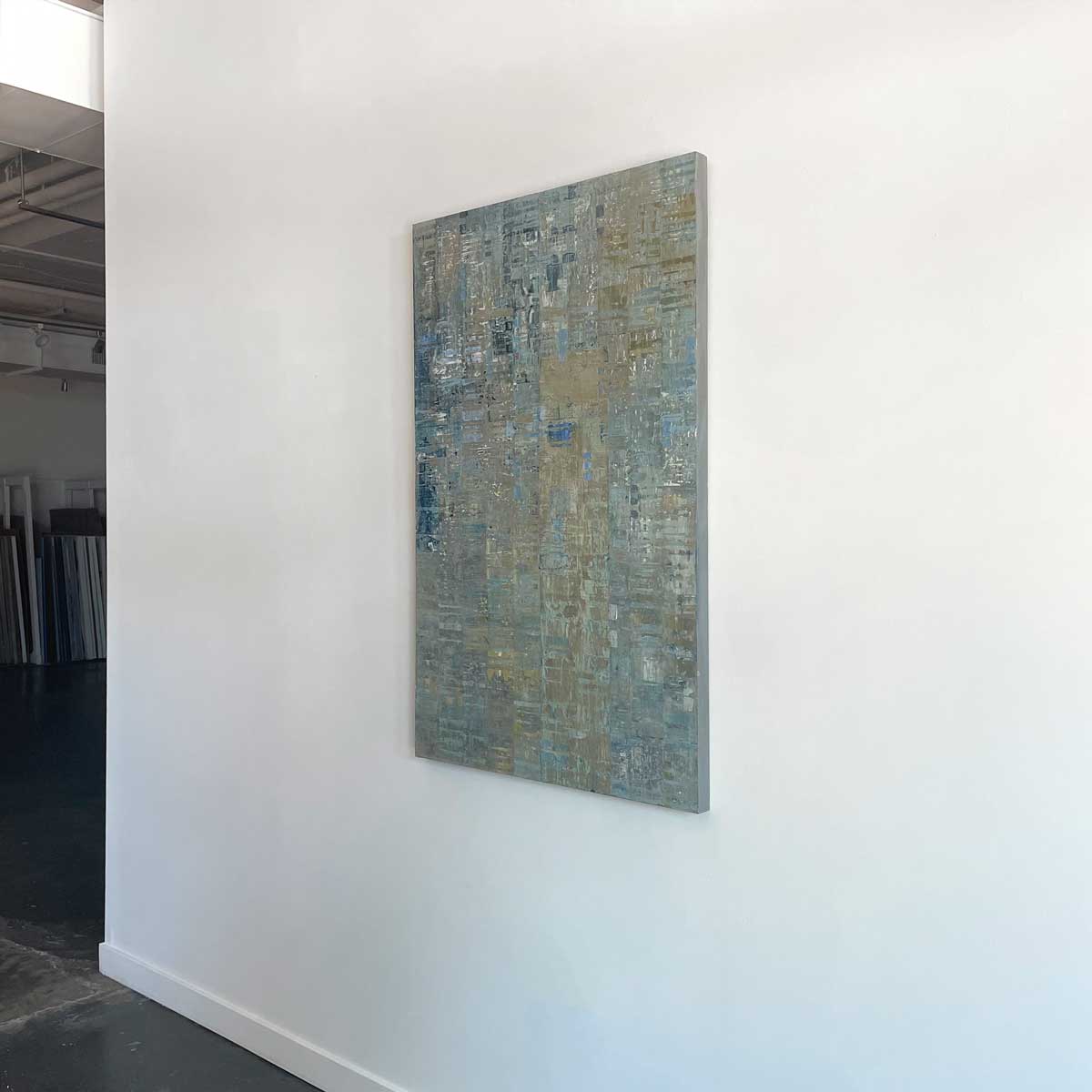 A blue and grey abstract painting with a silver leaf detail by Ned Martin hangs on a white wall with a view from the right. 