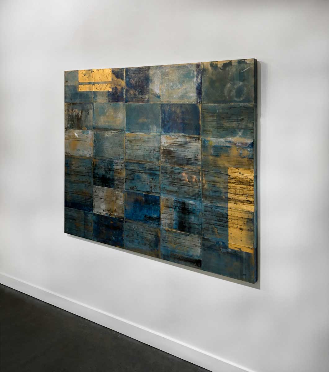 A right side-angle view of a blue, light blue and gold geometric abstract painting hangs on a white wall.