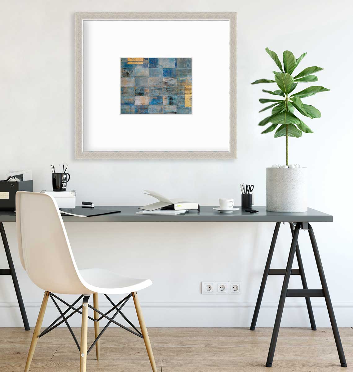 A blue and gold geometric abstract print in a silver frame with a mat hangs on a white wall above a grey desk with open notebooks, coffee mugs and a potted plant.