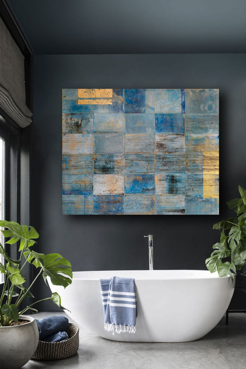 A blue, light blue and gold geometric abstract painting hangs on a charcoal wall above a white bathtub with a blue and white towel draped over the side, a woven basket with blue towels and a potted floor plant.