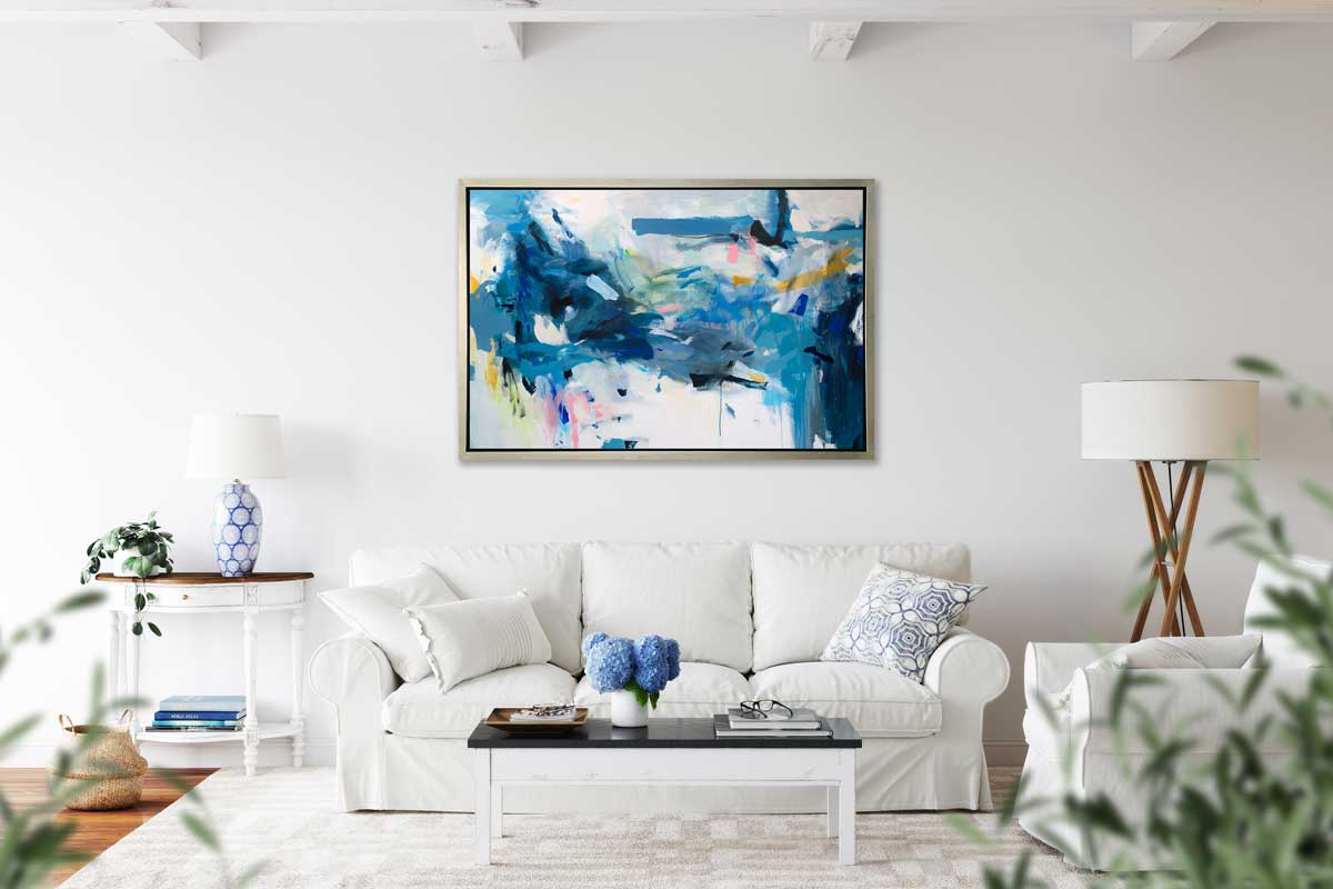 A blue, yellow and white abstract print in a silver floater frame hangs on a white wall above a white sofa with pillows, a black coffee table, a half-circle console table with a lamp and a floor lamp.