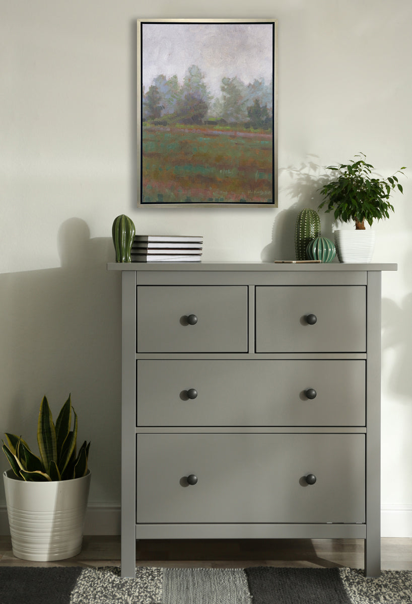 A grey and muted green abstract landscape print in a silver floater frame hangs on a white wall above a grey dresser with small potted plant, a stack of books and a few green cacti decorations.