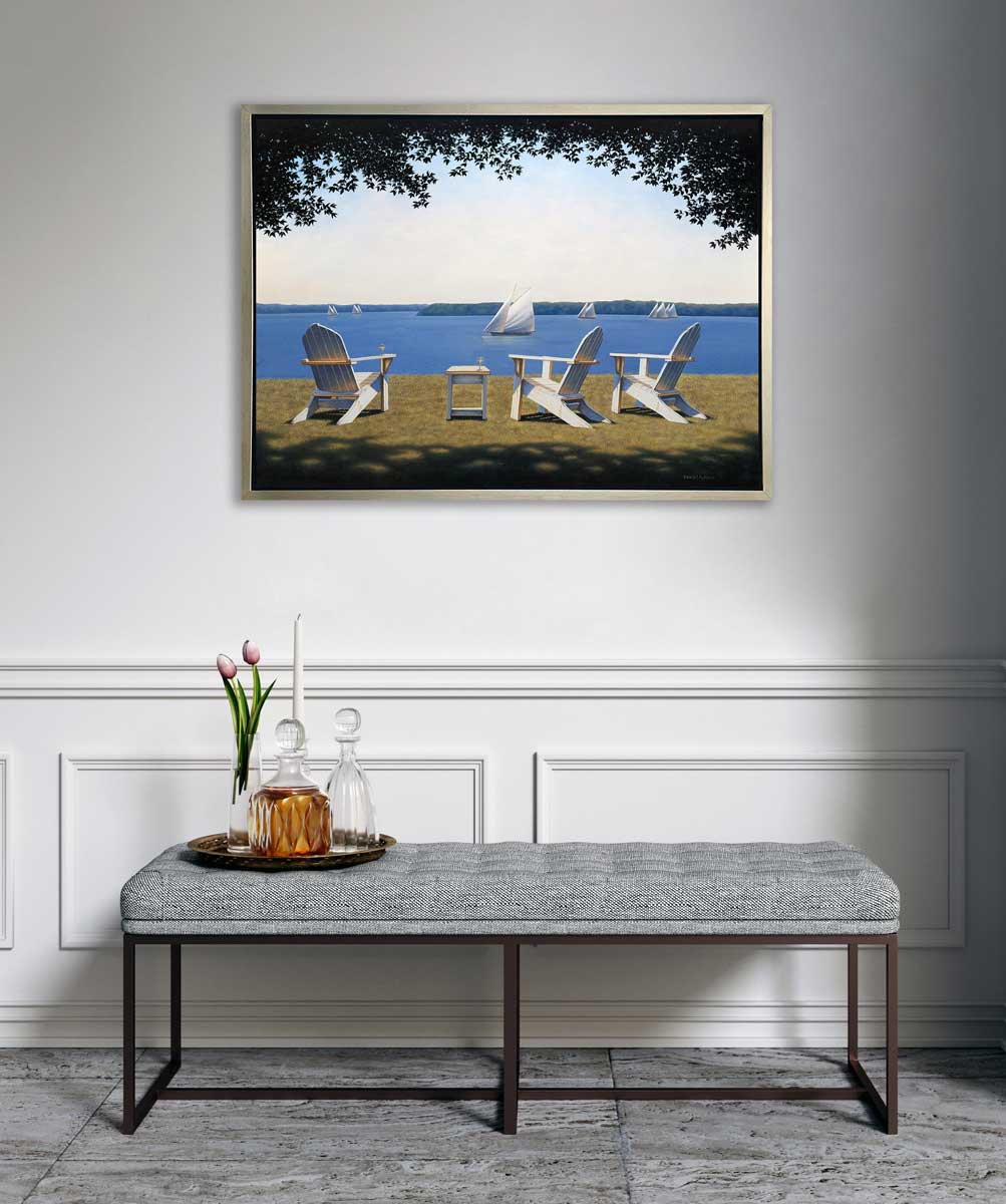 A realistic seascape with adirondack chairs print in a silver floater frame hangs on a white wall above a grey tufted bench with a tray of two decanters, a white tapered candlestick and a clear vase with two pink tulips.