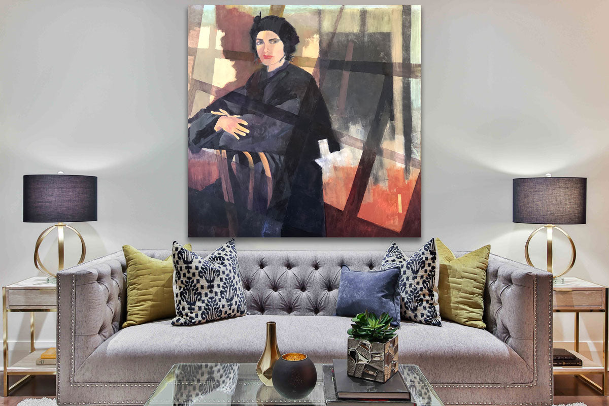 A large painting of a woman sitting backwards in a chair hanging over a gray tufted sofa with yellow and blue throw pillows. An end table and modern lamp are situated on each side of the sofa.