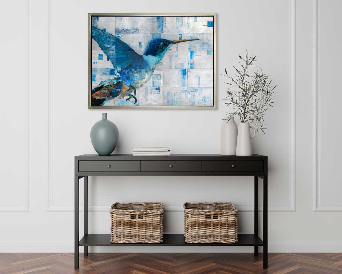 A blue and silver geometric abstract print of a hummingbird in a silver floater frame hangs on a white wall above a black console table with a muted teal vase, two white vases with greenery and two woven baskets on the shelf below.