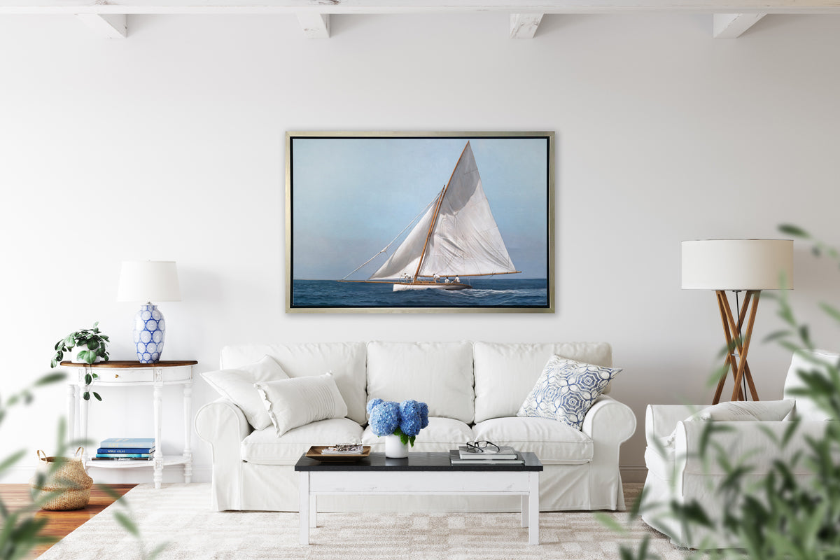 A blue, white and tan contemporary seascape print with a sailboat in a silver floater frame hangs on a white wall above a white sofa with pillows, a white and black coffee table, a white armchair and a tall floor lamp.
