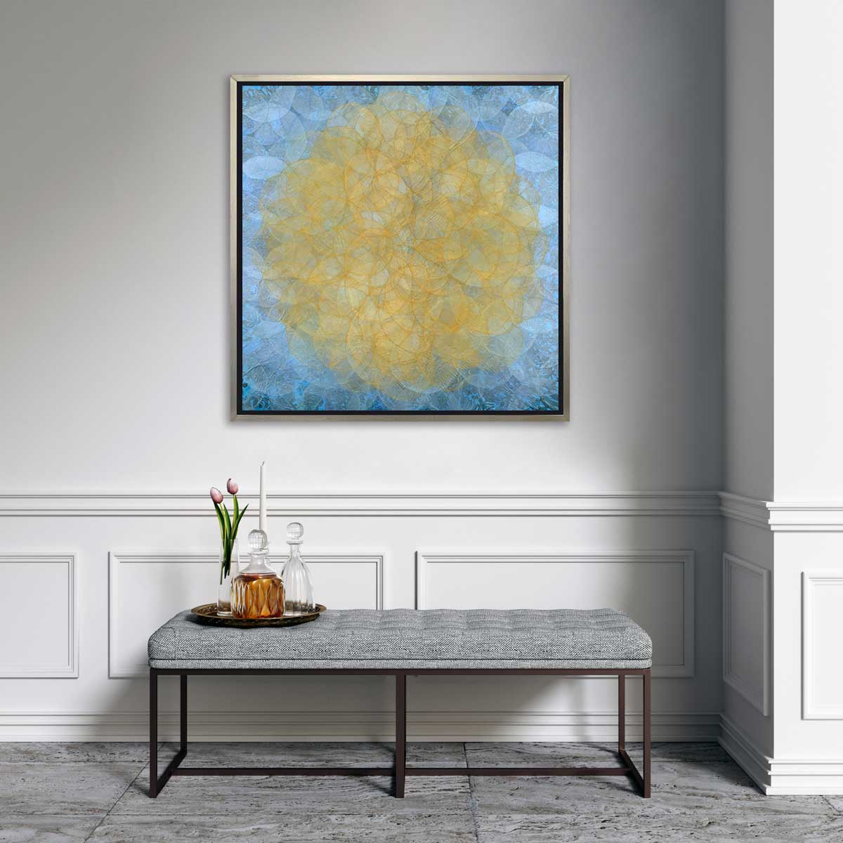 A blue, golden yellow and white abstract geometric print in a silver floater frame hangs on a white wall above a grey tufted bench with a tray filled with two glass decanters, a white tapered candle and a glass vase with two pink tulips.