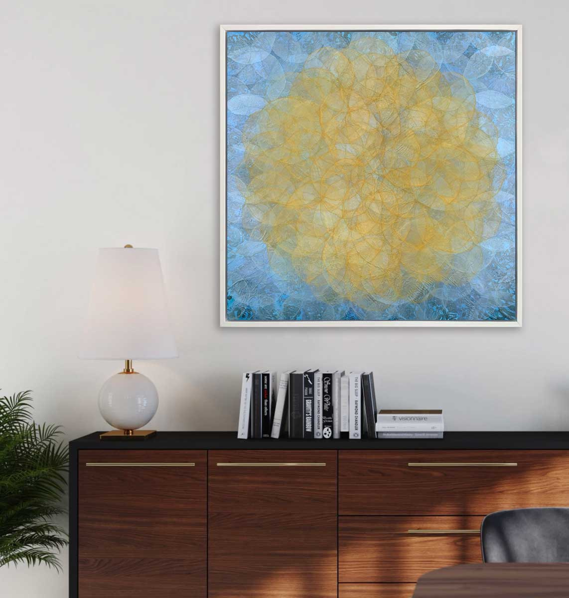 A blue, golden yellow and white abstract geometric print in a white floater frame hangs on a white wall above a black and wood credenza with a small white lamp and a stack of books.