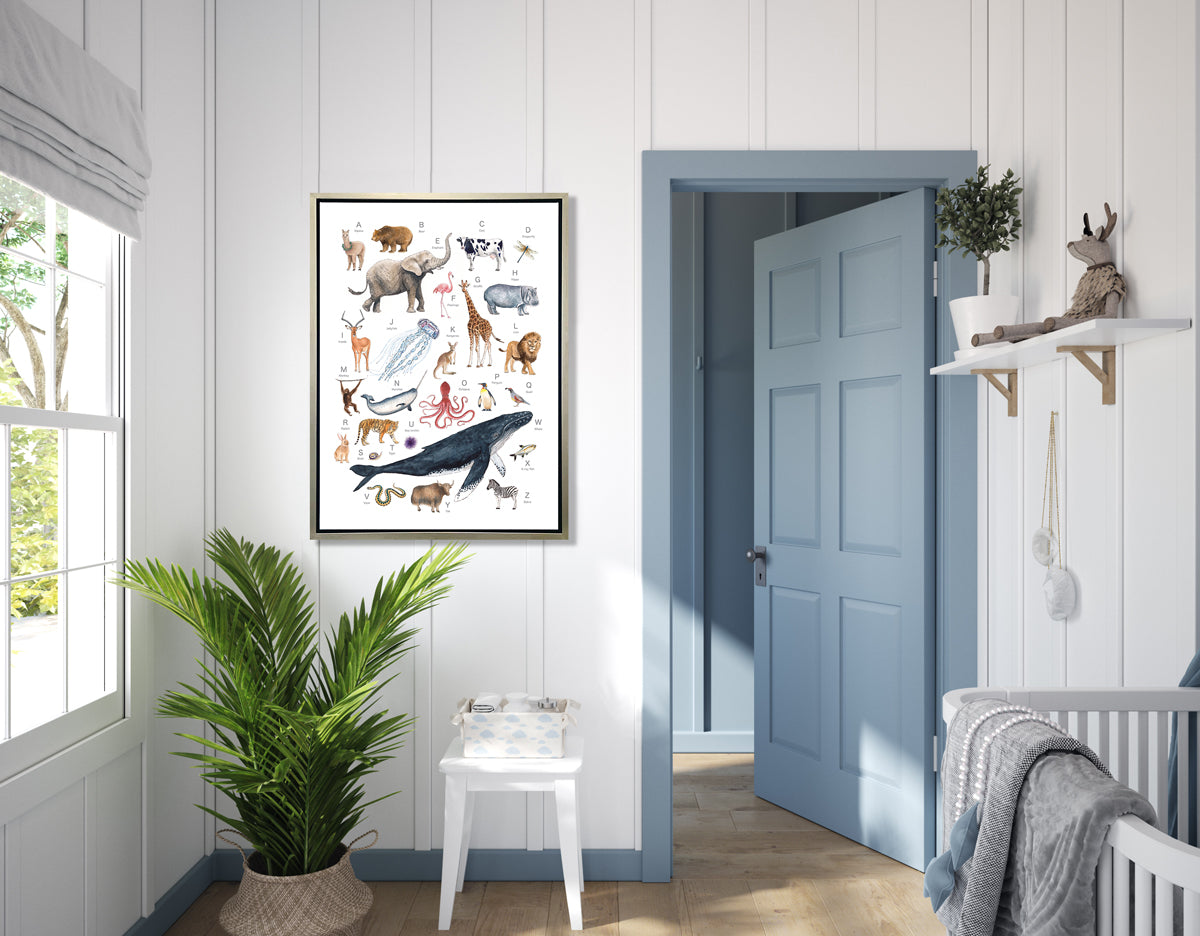 A multicolored print of different animals through the alphabet in a silver floater frame hangs on a white wall above a potted floor plant, a white side table with a baby basket and next to a blue door and a white crib.
