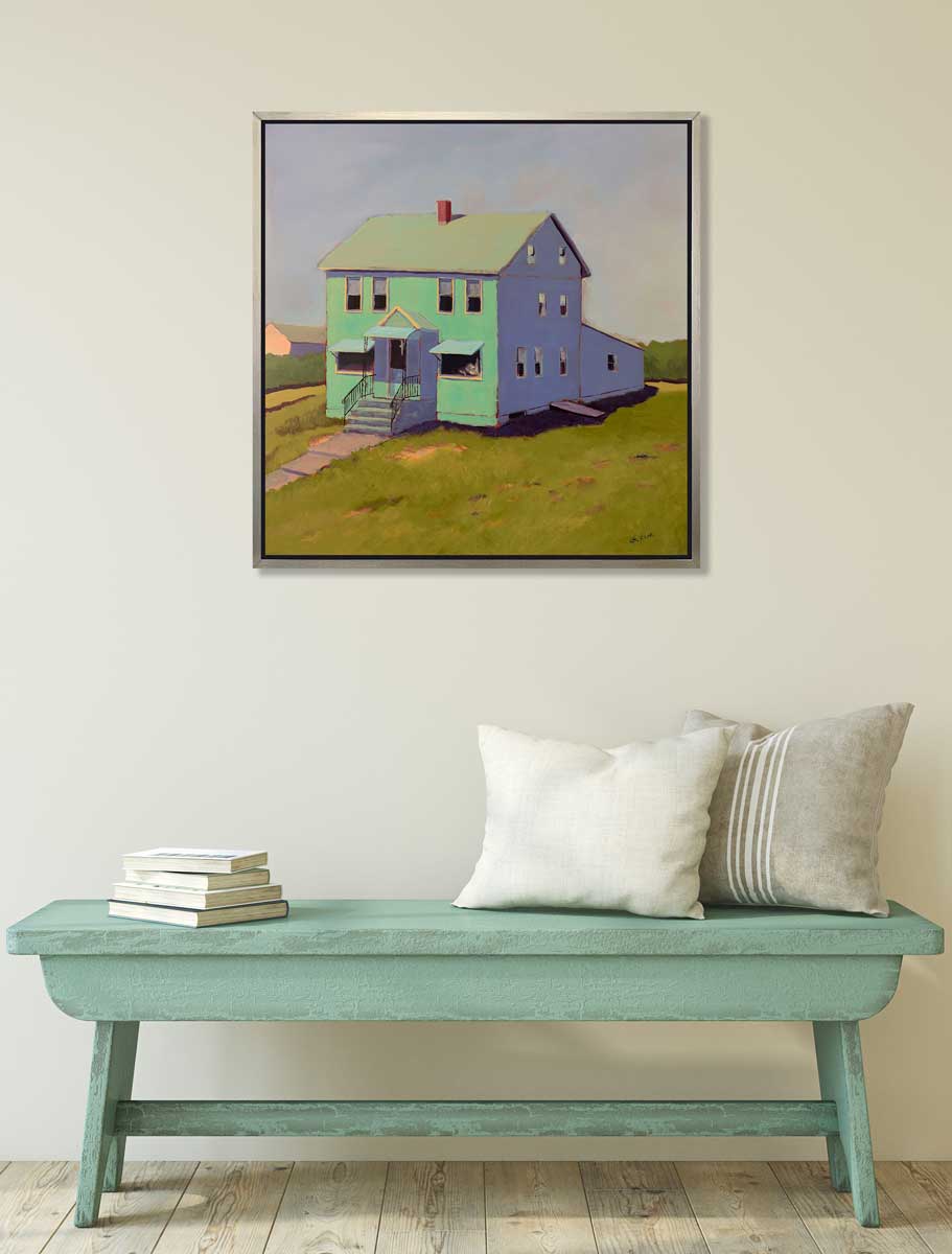 A seafoam green, blue and chartruse contemporary house print in a silver floater frame hangs on a white wall above a teal bench with a stack of books and two pillows.