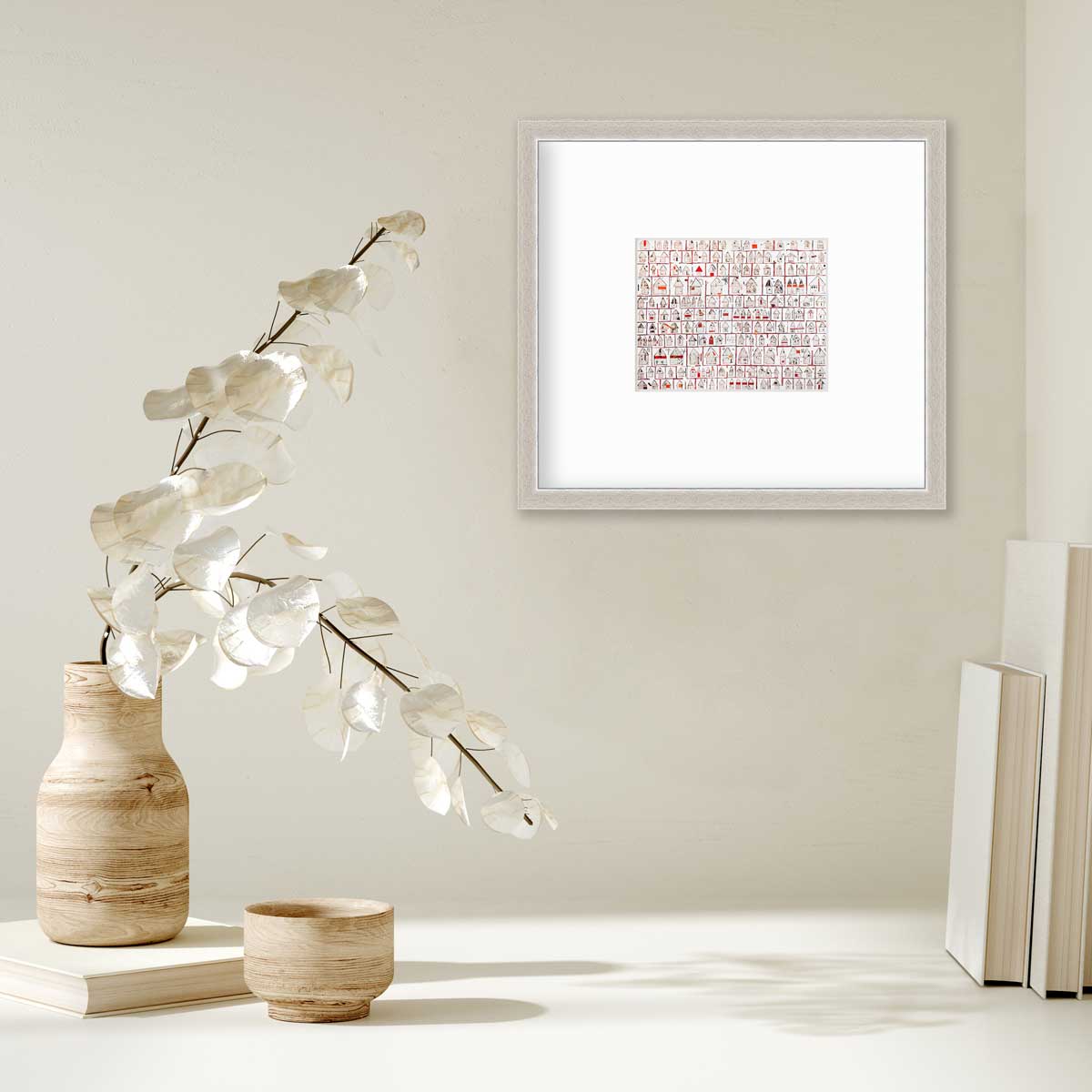 A red and white abstract house print in a silver frame with a mat hangs on a white wall above a stack of books and a wooden vase with white leaf stems.