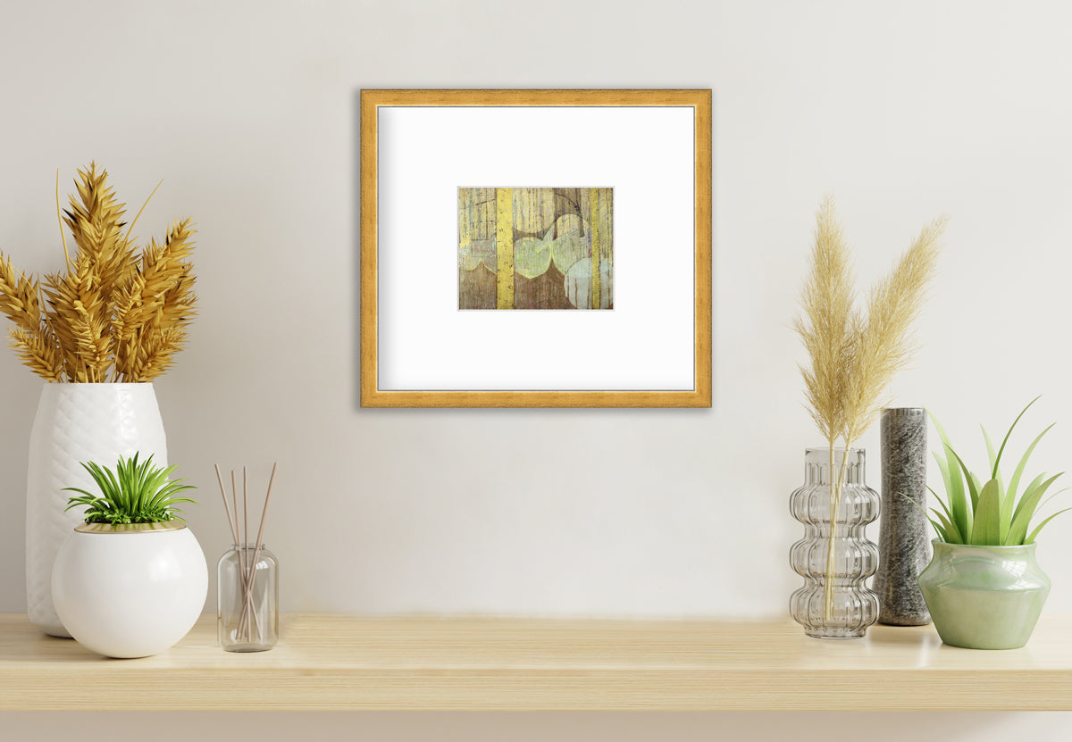 A gold, yellow, light green and light blue abstract in a gold frame with a mat hangs on a white wall above a wood surface with several vases full of greenery and wheat stems.