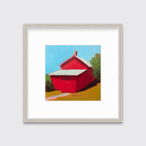 A print of a red barn with a grey roof, blue sky, green bushes and a chartreuse lawn in a silver frame with a mat hangs on a white wall.
