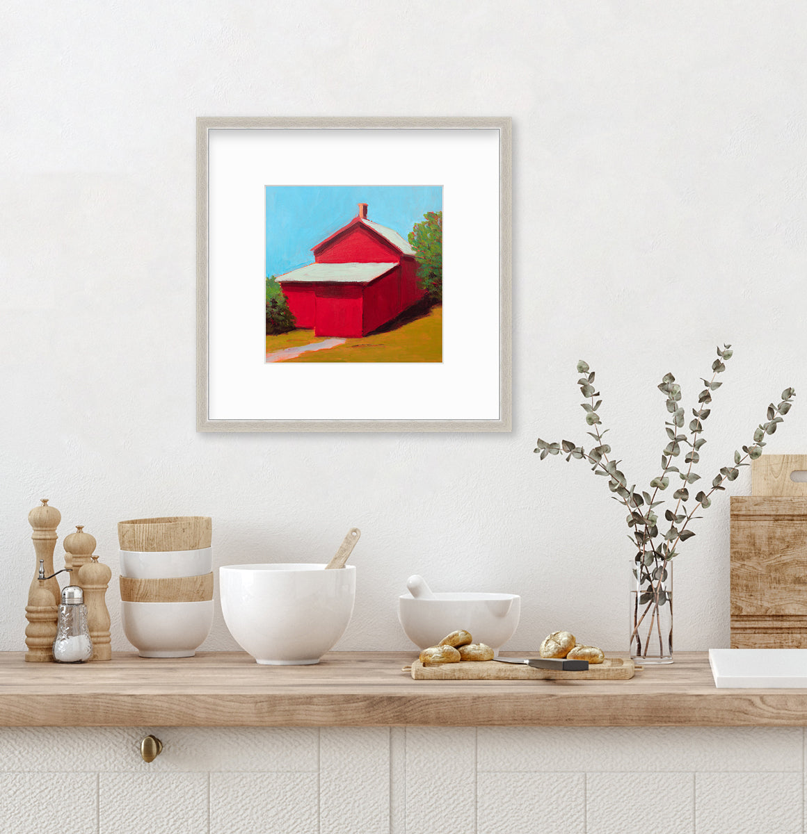 A print of a red barn with a grey roof, blue sky, green bushes and a chartreuse lawn in a silver frame with a mat hangs on a white wall above a wood countertop with a stack of bowls, a cutting board and a glass vase with eucalyptus stems.