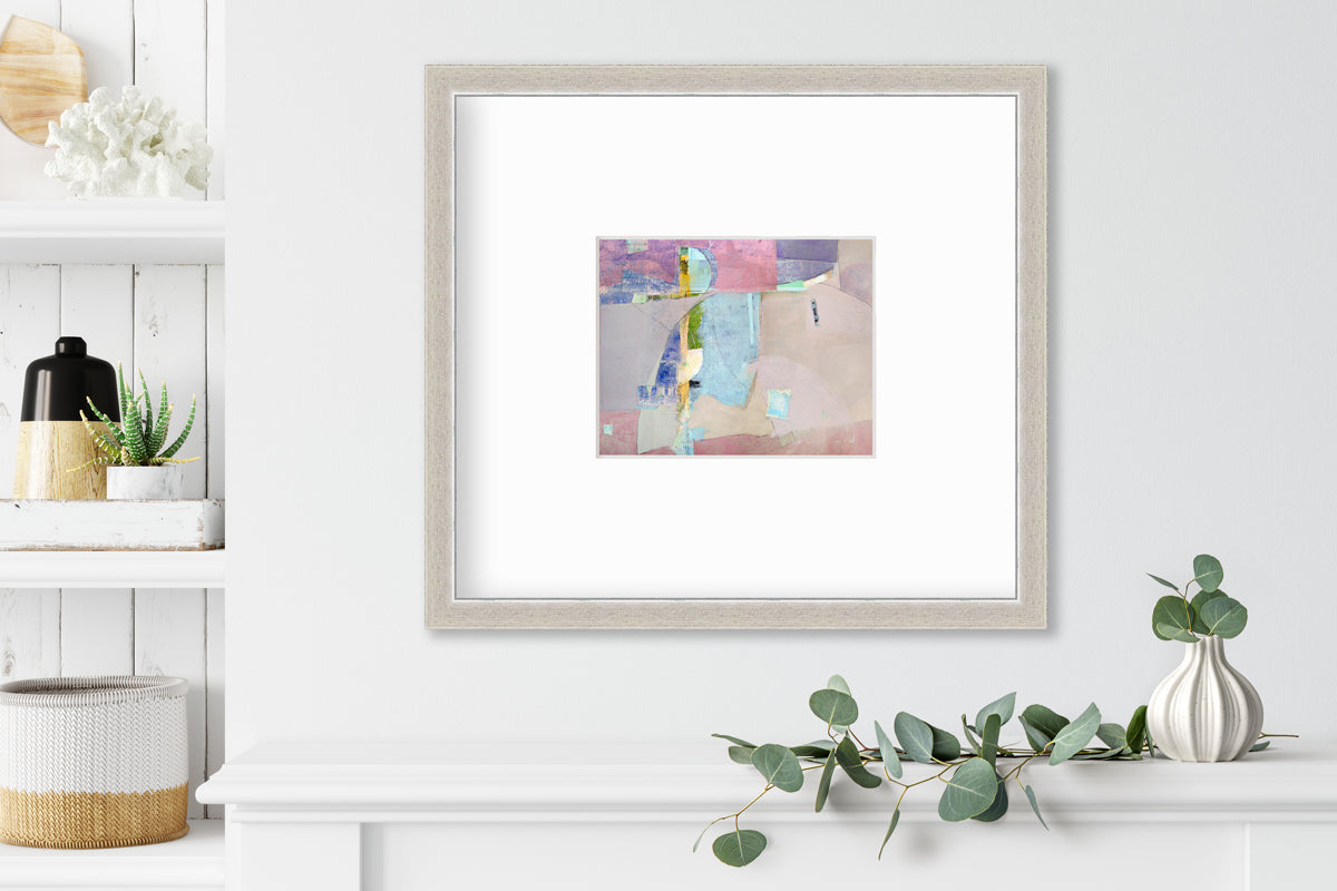 A pink, light blue, green and purple abstract print in a silver frame with a mat hangs on a white wall above a white shelf with a eucalyptus filled white vase and next to a built in bookshelf with decorative accessories.