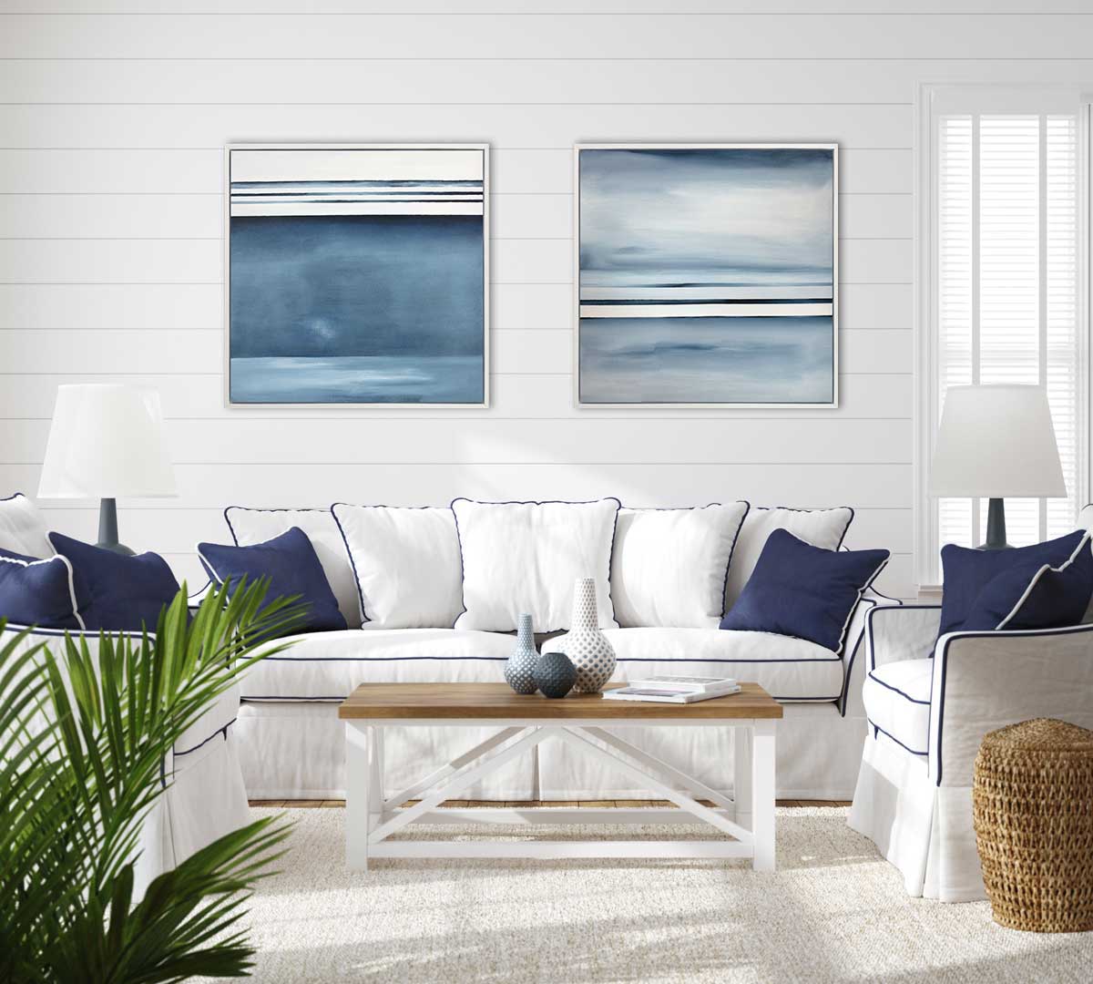 Two blue and white abstract painting in a white floater frame hanging on a white panel wall above a white sofa and two arm chairs with a blue trim.